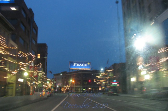 Photo of downtown with a billboard for peace. The street is lined with blurred Christmas lights. 