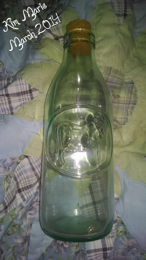 A glass bottle on a quilted bed spread with lavenders, greens, and blues. 