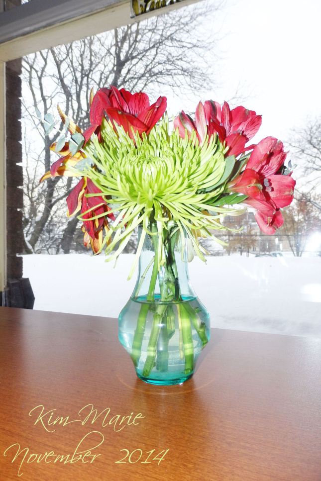 Some red flowers with a large green one in the middle. No, I don't know the names, just what looks pretty to me. They are in a vase on a desk facing out of a window onto a snowy, flat rooftop. 