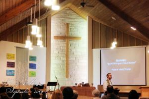 Church santuary with pastor preaching, the cross on the wall, and the backs of people's heads in the pews.