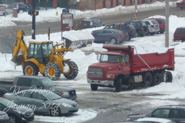 A front end loader dumping snow into the bed of a truck to haul the snow out of a parking lot.