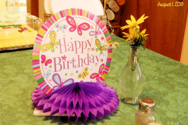 The image of a table center piece made of round paper with a pink stripped border and butterflies wishing a Happy Birthday. Purpose tissue stuff below. Yellow field flowers in a small vase beside it.
