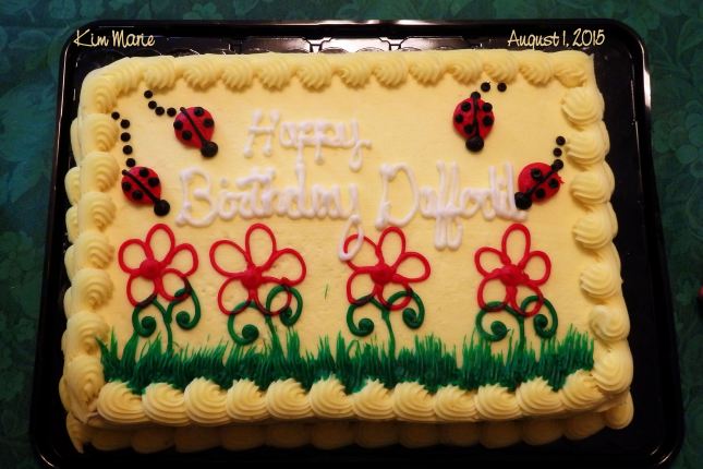 A yellow birthday cake with gree grass, red flowers, lady bugs, and a greeting,