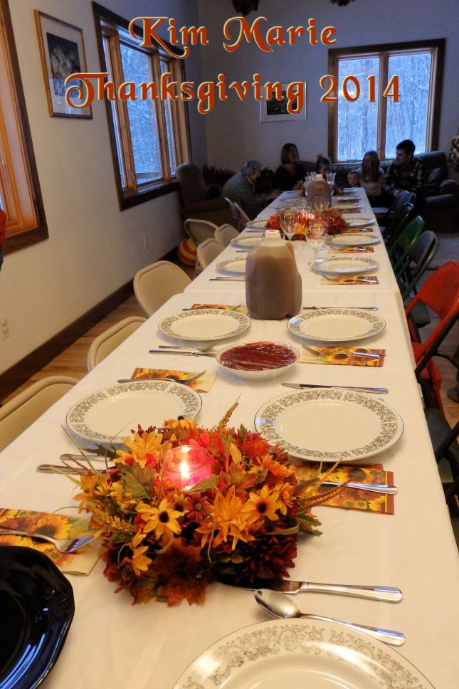 The table is set for Thanksgiving with china plates, silverware, autumn leaf napkins, a floral arrangement with a candle in the center, a gallon of cider, and quite the long, long table.