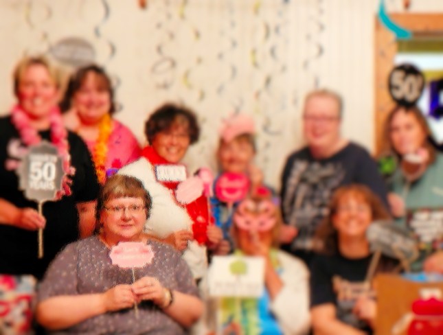 9 women celebrating a couple of 50th birthdays. Most dressed in black or pink, but a few in gray or blue. Most holding a 50th sign of some sort. Streamers hanging from the ceiling.