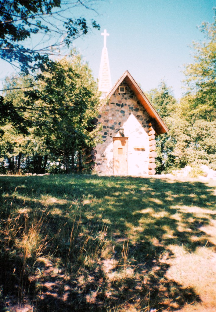 A grassy hill, with trees at the top, and a small stone chapel in the middle of the trees. The stone chapel has a wood door, no windows that I can see (although I thought there were form my memories, but I could remember wrong). There is a tall steeple with a small white cross on the top of the roof of the chapel. The trees are shading the hillside. 