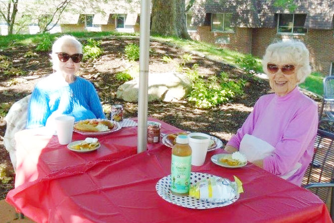 Two women of a certain age (numerous decades) are sitting at a table with a red table covering, with plates of lunch in front of them, wearing sunglasses, and smiling as I take their photo.
